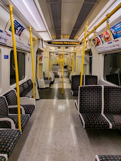 Close-up view of the exterior of several London Underground train carriages parked at a platform, with the train doors visible and closed. The platform surface features a tactile yellow and black stri