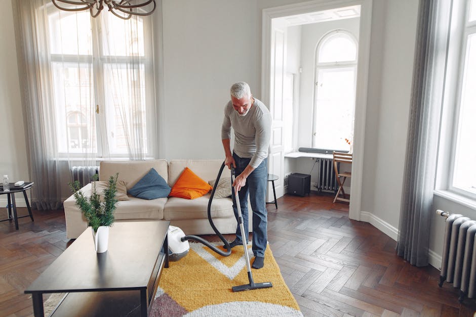 A professional cleaner from Bermondsey Carpet Cleaners is performing surface cleaning in a bright living room. The cleaner, an older man with gray hair and glasses, is vacuuming a yellow patterned rug
